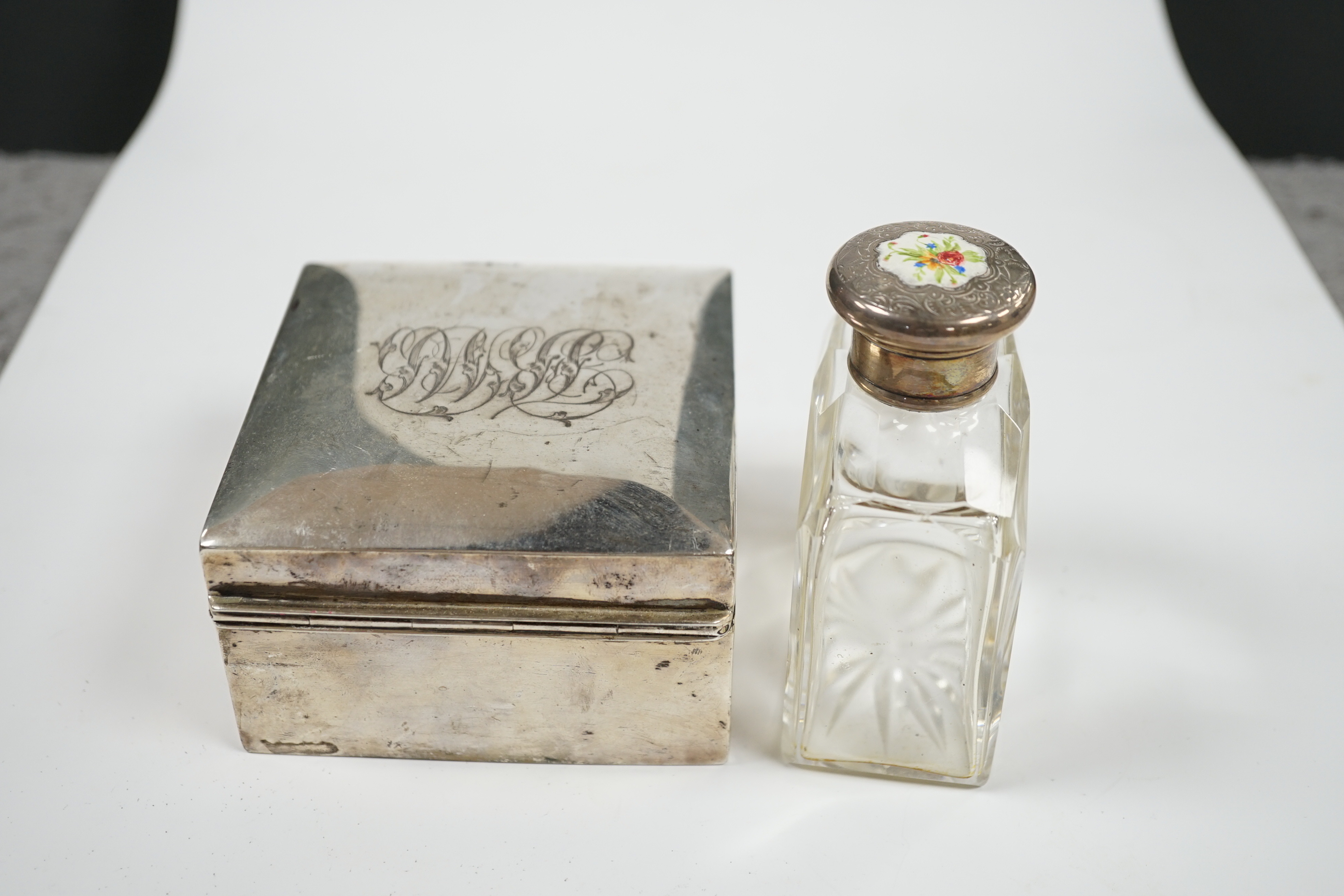 An Edwardian silver mounted cigarette box, marks rubbed, 90mm, together with a white metal and enamel topped glass scent bottle. Condition - poor